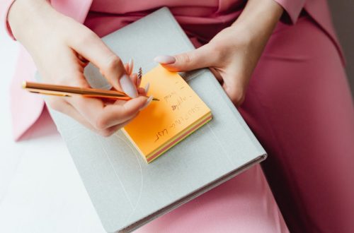Close-up of a woman writing on a sticky note with a pencil, wearing a pink suit.